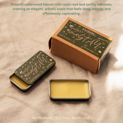 A small rectangular metal tin of balm with pale yellow wax sits in front of its kraft paper box and matching lid, all printed with the scent name "Hyunjin's Lovestay"and brand "Puchhka". The products are arranged on a beige fabric background with descriptive text at the top and bottom.