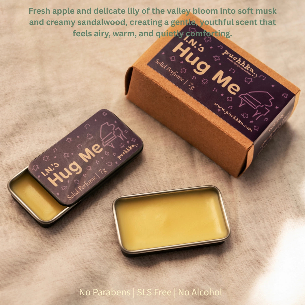 Solid perfume balm tins and packaging on a textured beige surface. One tin is open, showing pale yellow balm, one tin is closed with a purple label reading 'I.n's Hug Me', and the product box is brown kraft paper with a matching purple label.