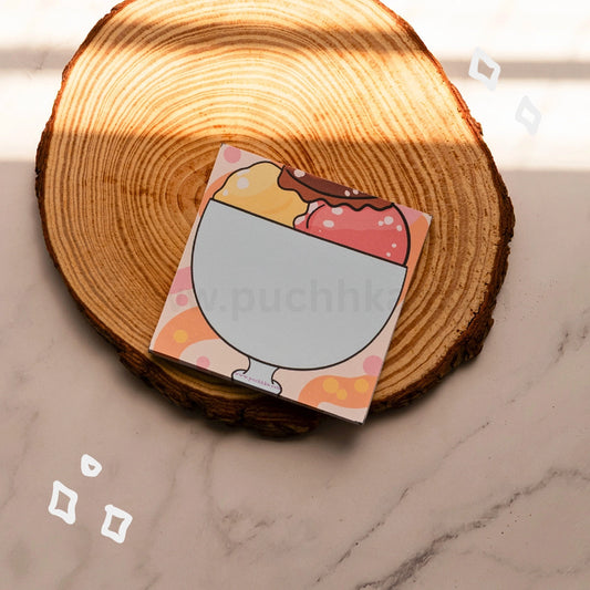 A square product box with an illustration of a dessert glass sits on a round wood  coaster over a light marble surface. 