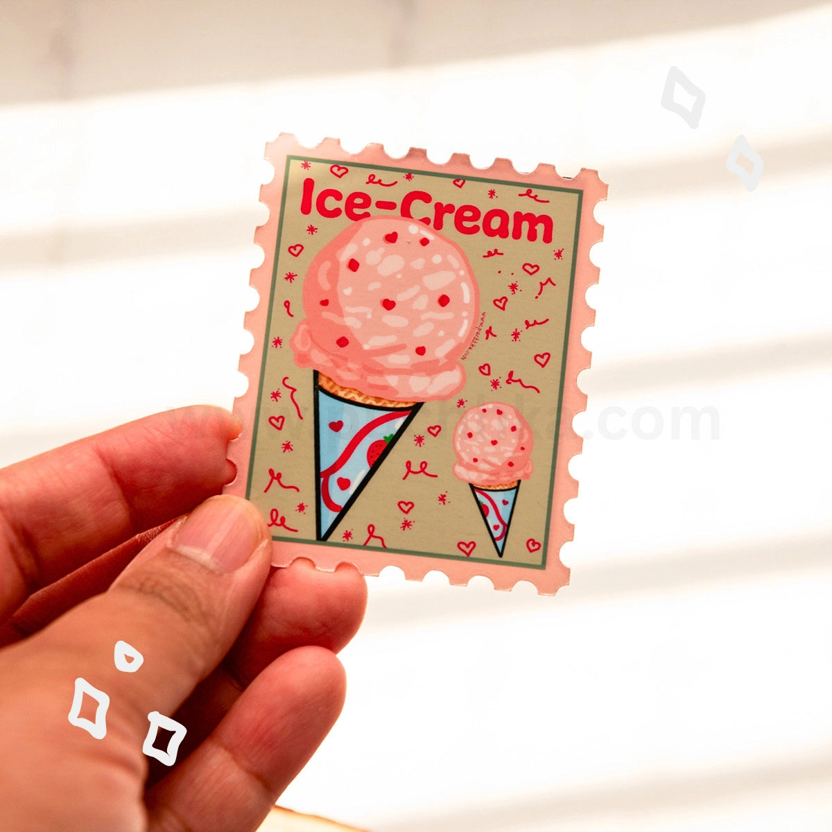 A hand holds a postage-stamp-style sticker featuring pink ice cream scoops in cones on a beige background with the words “Ice-Cream” at the top.