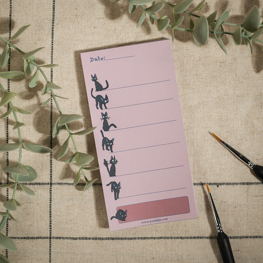 A pink notepad with seven illustrated cat poses and a date line, styled on a beige fabric surface with green eucalyptus leaves and two paintbrushes.