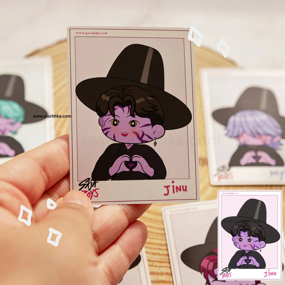 A hand holds a printed card featuring a chibi-style illustration of a person in a black witch hat and black outfit with purple skin tones and a pink heart. Other similar cards are visible in the background on a light wood surface.