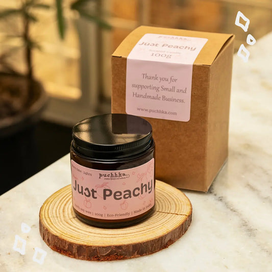 A jar of Just peachy scented candle  with a black lid and pink label reading 'Just Peachy' sits on a round wood coaster, with its kraft paper box behind it on a marble surface.