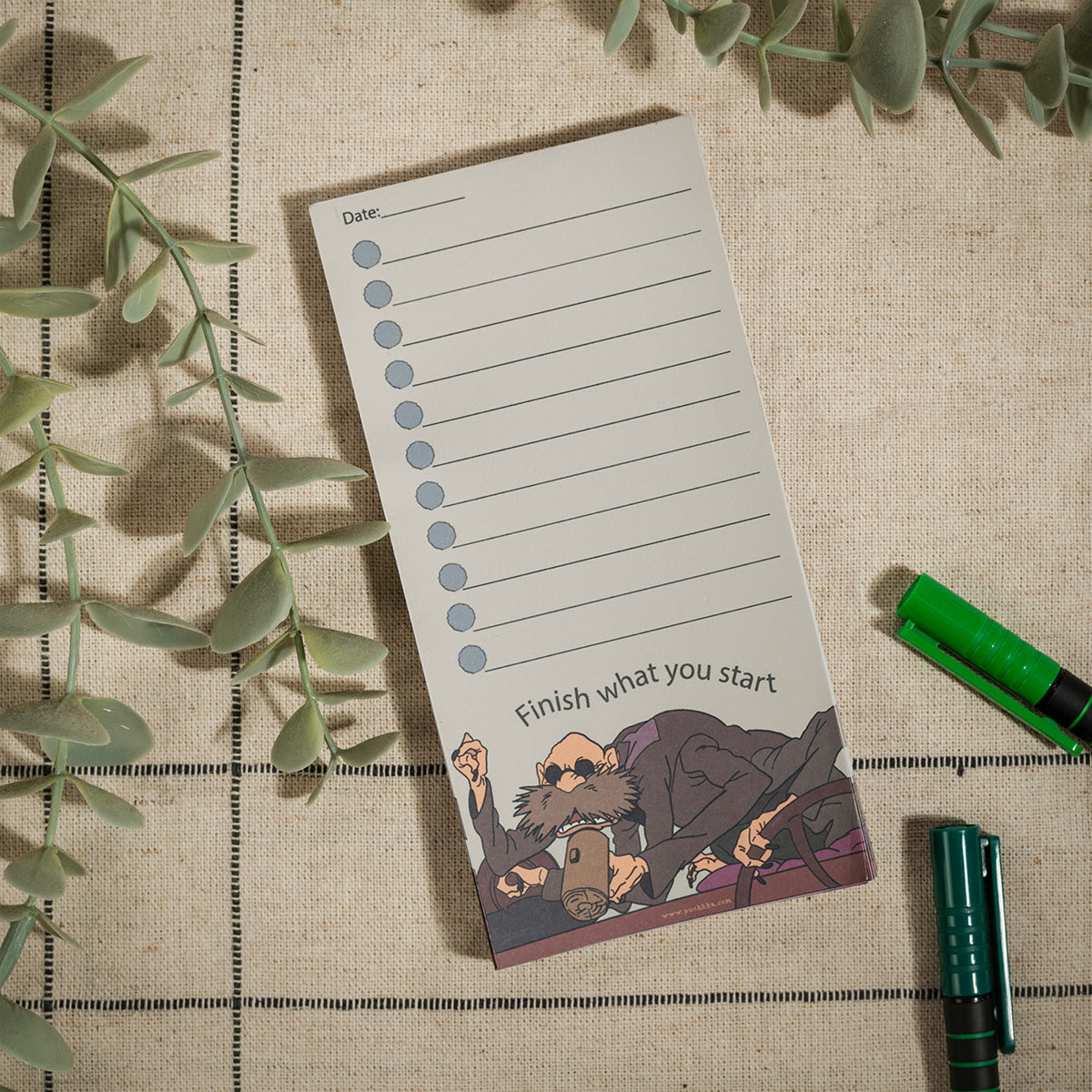 A vertical notepad with lined writing space, a blue circle checklist, and the phrase 'Finish what you start' at the bottom, styled on a beige fabric surface with greenery and pens.
