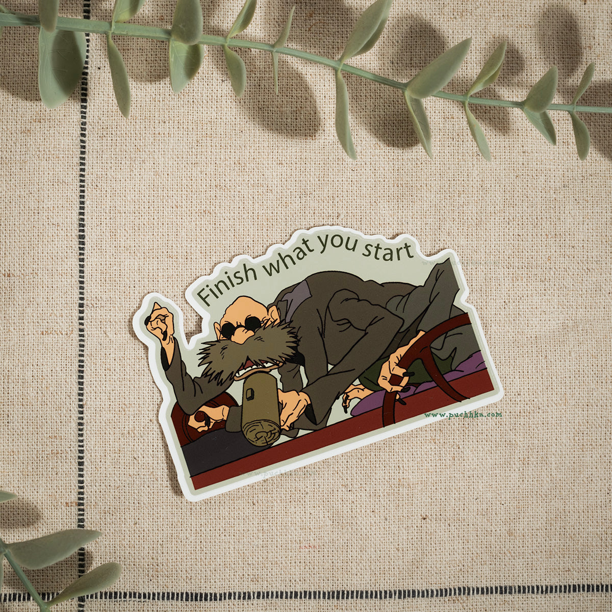 A die-cut sticker featuring an illustrated bearded man in a suit holding a mug, with the text 'Finish what you start' above him, placed on a beige textured fabric background with green leaves around the edges.
