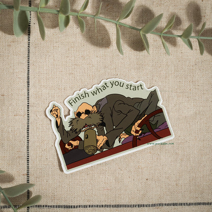 A die-cut sticker featuring an illustrated bearded man in a suit holding a mug, with the text 'Finish what you start' above him, placed on a beige textured fabric background with green leaves around the edges.