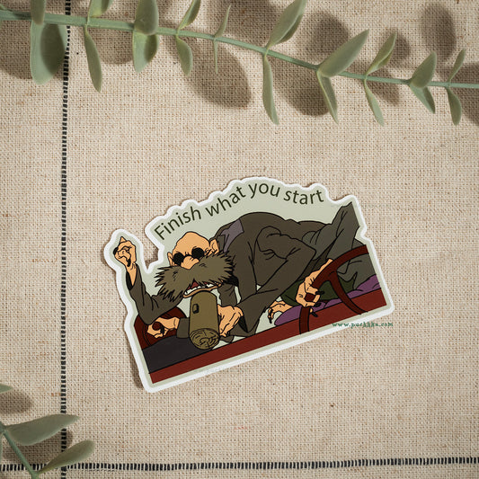 A die-cut sticker featuring an illustrated bearded man in a suit holding a mug, with the text 'Finish what you start' above him, placed on a beige textured fabric background with green leaves around the edges.