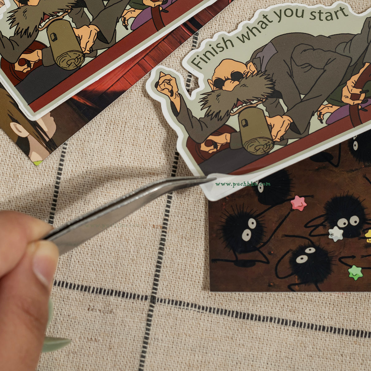 A hand uses metal tweezers to place a die-cut sticker featuring a cartoon bearded man with the text 'Finish what you start' onto a printed card. Other stickers are visible in the background on a textured beige fabric surface.