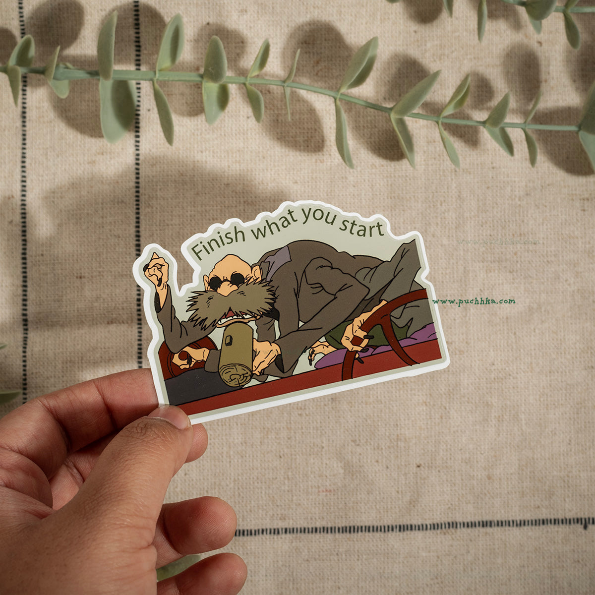 A hand holds a die-cut sticker featuring a cartoon illustration of a bearded man in sunglasses lounging on a couch with the text 'Finish what you start'. The sticker is shown against a beige linen background with green eucalyptus leaves.