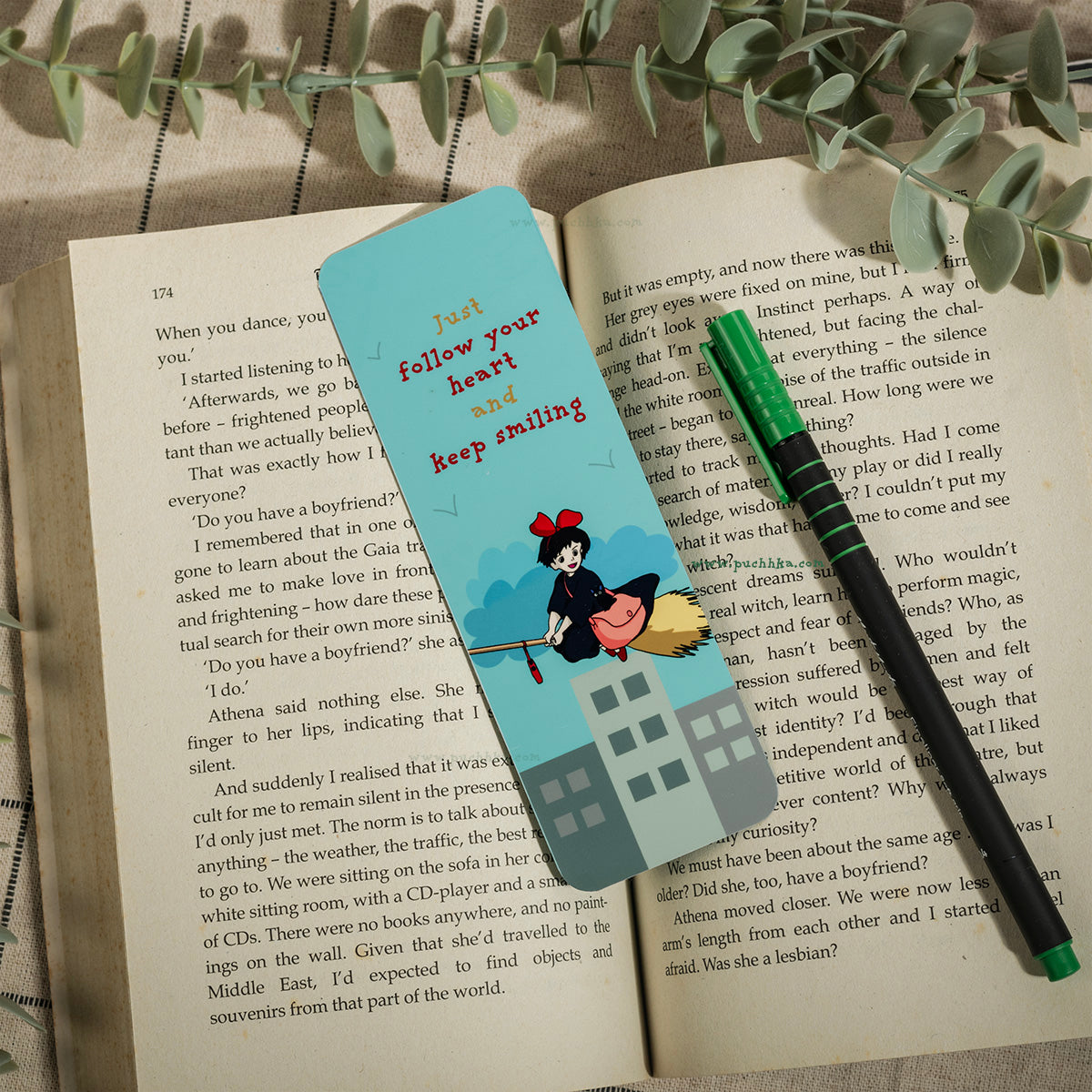 A light blue rectangular bookmark with a ghibli character and the words 'Just follow your heart and keep smiling' lies on top of an open book. A green pen rests on the right side of the bookmark.
