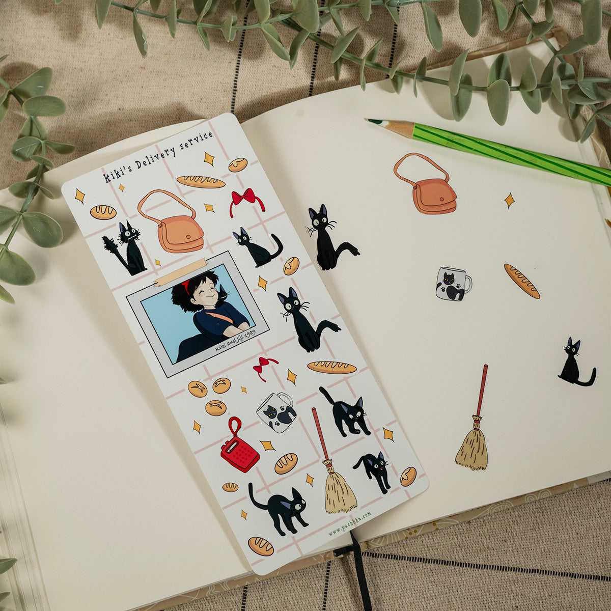 A notebook page decorated with cute anime-style stickers, including black cats, bread loaves, handbags, and a character portrait, styled on a neutral fabric background with a green pencil and eucalyptus leaves.