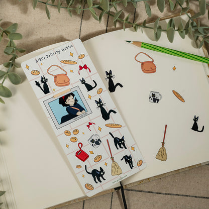 A notebook page decorated with cute anime-style stickers, including black cats, bread loaves, handbags, and a character portrait, styled on a neutral fabric background with a green pencil and eucalyptus leaves.