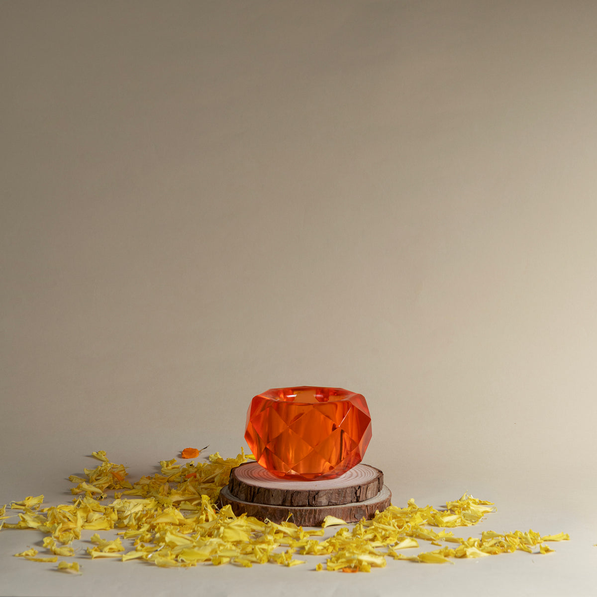 A faceted orange resin bowl or candle holder sits on two stacked wood slices, surrounded by scattered yellow petals against a plain beige studio background.