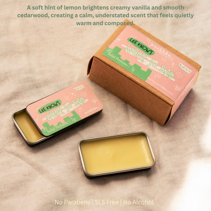 Lee Know's 'Want So Bad' solid perfume shown in its packaging, featuring a brown cardboard box and two small metal tin containers with pink labels, arranged on a beige fabric background with descriptive text overlays.
