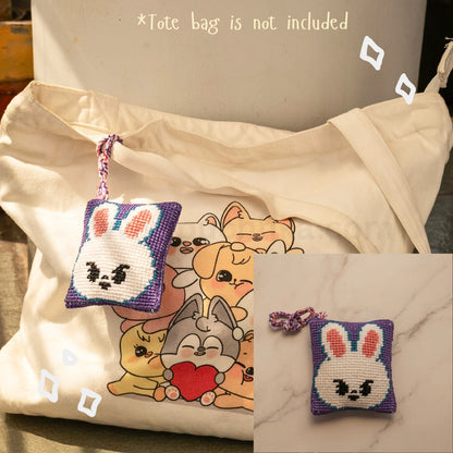 A cream canvas tote bag with a printed skzoo illustration on the front, shown alongside a small lavender and white cross-stitch style skzoo bag hanging with a matching strap. The image includes text stating '*Tote bag is not included' and a small inset showing the pouch alone on a marble surface.
