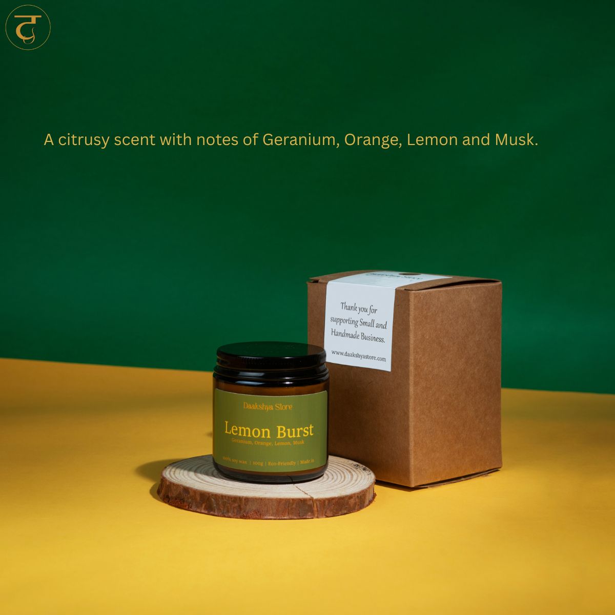 A candle jar labeled 'Lemon Burst' with a black lid and green label, placed on a round wood coaster next to its brown kraft box. The scene is set against a split green and yellow background with text describing the citrus scent.