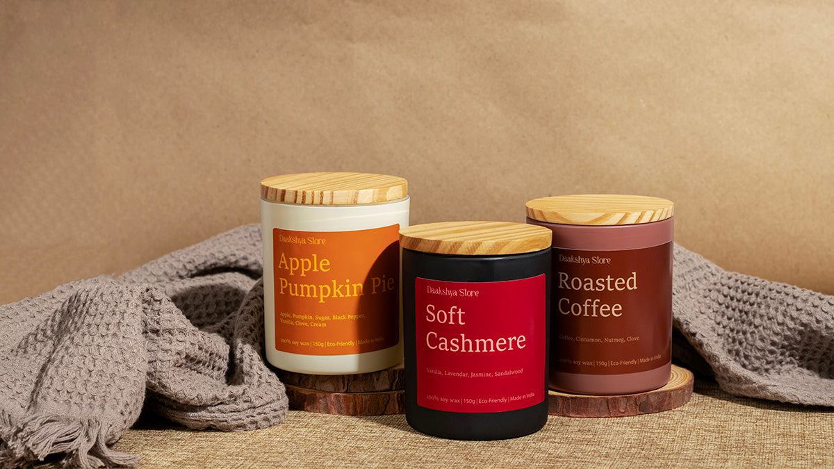 Three scented candles in jars with wooden lids are arranged on a textured surface with a neutral backdrop and gray knit fabric. The labels read 'Daakshya Store' and show scents: Apple Pumpkin Pie, Soft Cashmere, and Roasted Coffee.