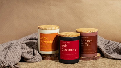 Three scented candles in jars with wooden lids are arranged on a textured surface with a neutral backdrop and gray knit fabric. The labels read 'Daakshya Store' and show scents: Apple Pumpkin Pie, Soft Cashmere, and Roasted Coffee.