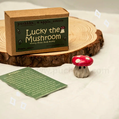 A small kraft paper box labeled 'Lucky the Mushroom' sits on a rustic wood coaster, with a printed card in front and a small red-and-white mushroom figurine beside it on a white surface.