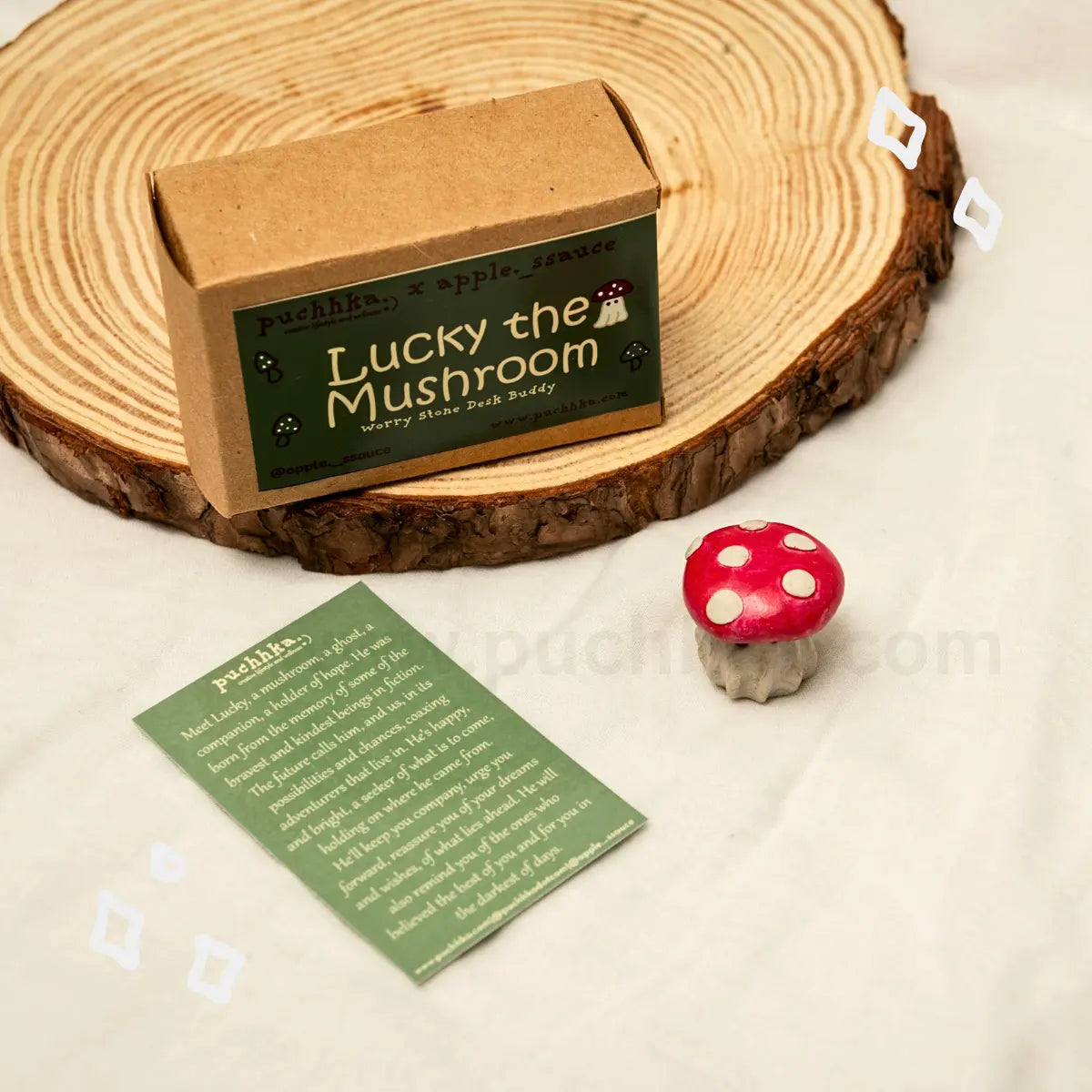 A kraft paper box labeled 'Lucky the Mushroom' sits on a round wood coaster, with a small red-and-white mushroom figurine and a green printed card on a light fabric background.