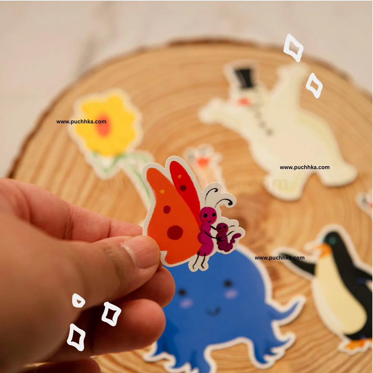 A hand holds a colorful butterfly sticker in front of a round wooden slice background, stickers blurred behind it. White website watermarks are visible across the photo.