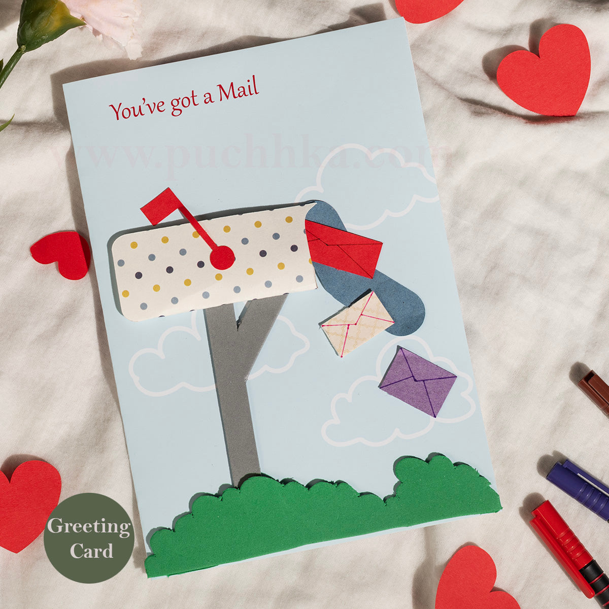 A handmade paper greeting card styled on a textured white fabric background with red paper hearts and markers. The card features a light blue sky background with a layered paper mailbox, green grass, and several colorful paper envelopes floating out.