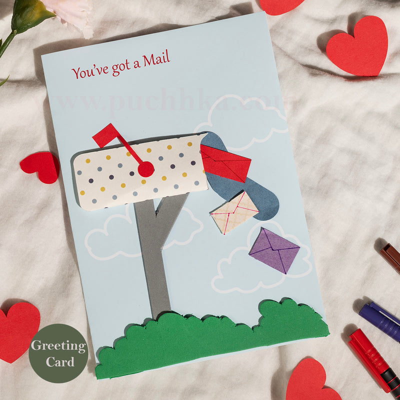 A handmade paper greeting card styled on a textured white fabric background with red paper hearts and markers. The card features a light blue sky background with a layered paper mailbox, green grass, and several colorful paper envelopes floating out.