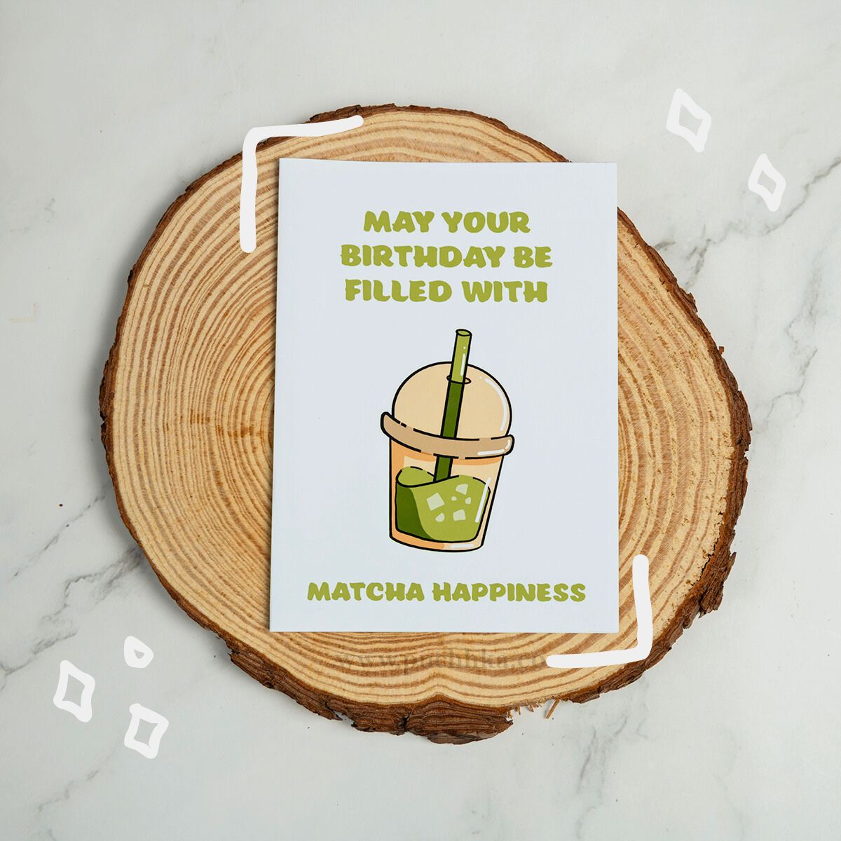 A square birthday card with a white background and green text reading 'MAY YOUR BIRTHDAY BE FILLED WITH MATCHA HAPPINESS,' featuring a matcha drink illustration in the center. The card is placed on a round wood slice coaster over a light marble surface.