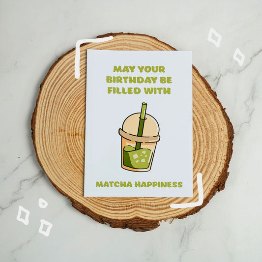 A square birthday card with a white background and green text reading 'MAY YOUR BIRTHDAY BE FILLED WITH MATCHA HAPPINESS,' featuring a matcha drink illustration in the center. The card is placed on a round wood slice coaster over a light marble surface.