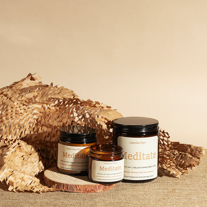 Three amber glass jars with cream labels and black lids, labeled 'Meditate' by Daakshya Store (now Puchhka), styled on a burlap surface with a wood coaster and woven natural paper against a beige background.