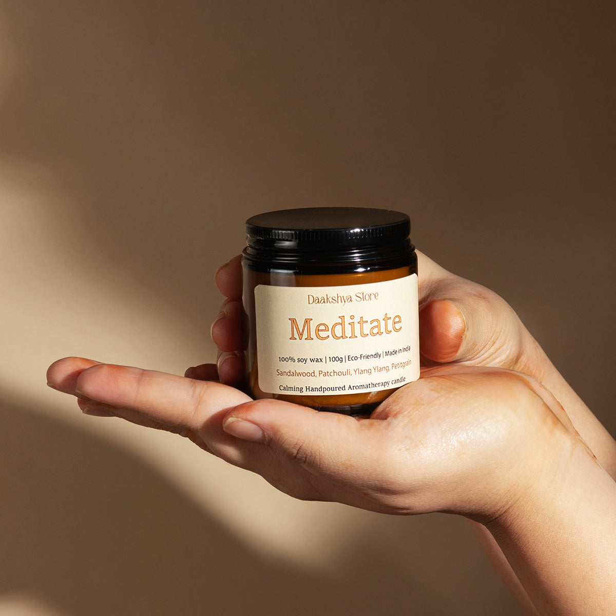 A hand holding a small amber glass jar labeled 'Meditate' by Daakshya Store (now puchhka), with a black lid and cream label, against a warm beige background with soft shadows.