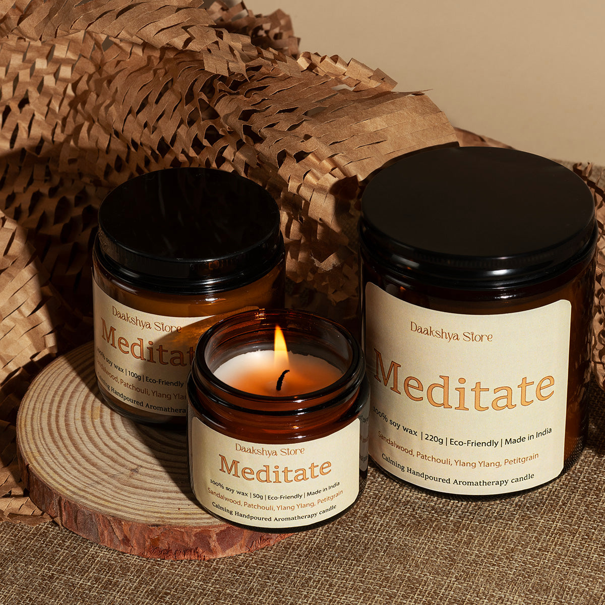 Three amber glass jars of Meditate scented candles from Daakshya Store, styled on a rustic wood slice with burlap and a woven basket background.