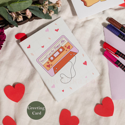 A square white greeting card featuring a retro cassette tape illustration with the phrase 'You and I are the Perfect Mix' and small pink hearts, styled on a cream fabric surface with red heart cutouts, flowers, and colored pencils.