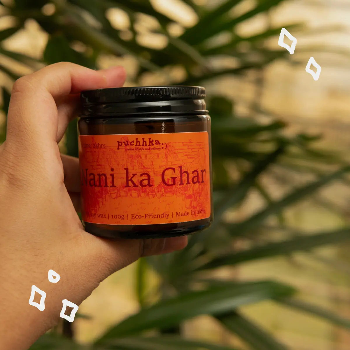 A hand holding a dark amber glass jar of Puchha 'Nani ka ghar' with a bright orange label and black lid, set against a blurred background of green leaves.