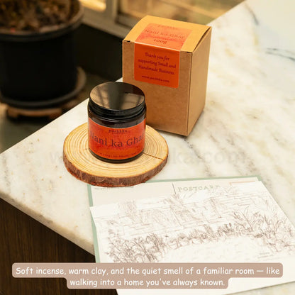 Nani ka Ghar scented candle on a wooden slice coaster next to its kraft paper packaging box on a white marble countertop. A printed postcard and a descriptive text overlay are in the foreground.