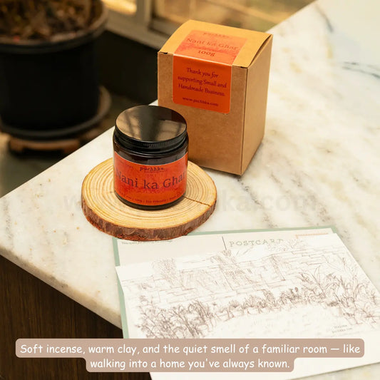 Nani ka Ghar scented candle on a wooden slice coaster next to its kraft paper packaging box on a white marble countertop. A printed postcard and a descriptive text overlay are in the foreground.