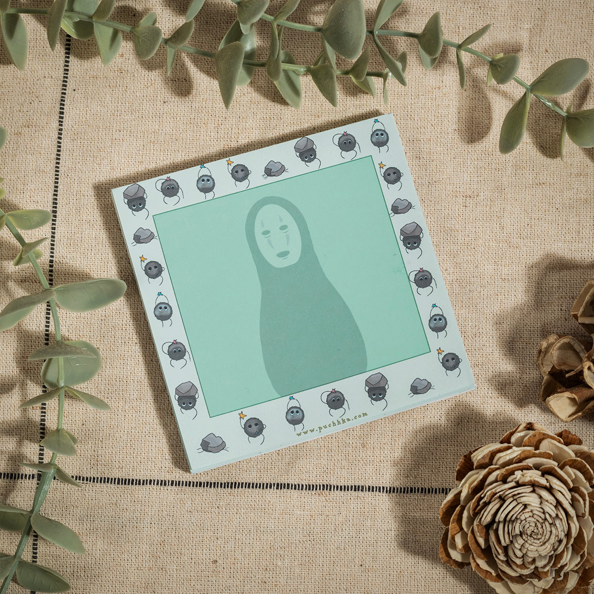A square art print featuring a stylized No-Face illustration on a mint green background, surrounded by a border of small soot sprites. The print is styled in a flat-lay on a beige linen fabric with green eucalyptus leaves and dried pinecones.