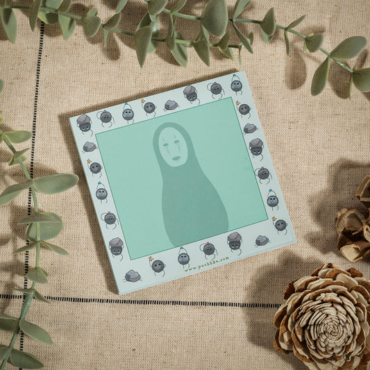 A square art print featuring a stylized No-Face illustration on a mint green background, surrounded by a border of small soot sprites. The print is styled in a flat-lay on a beige linen fabric with green eucalyptus leaves and dried pinecones.