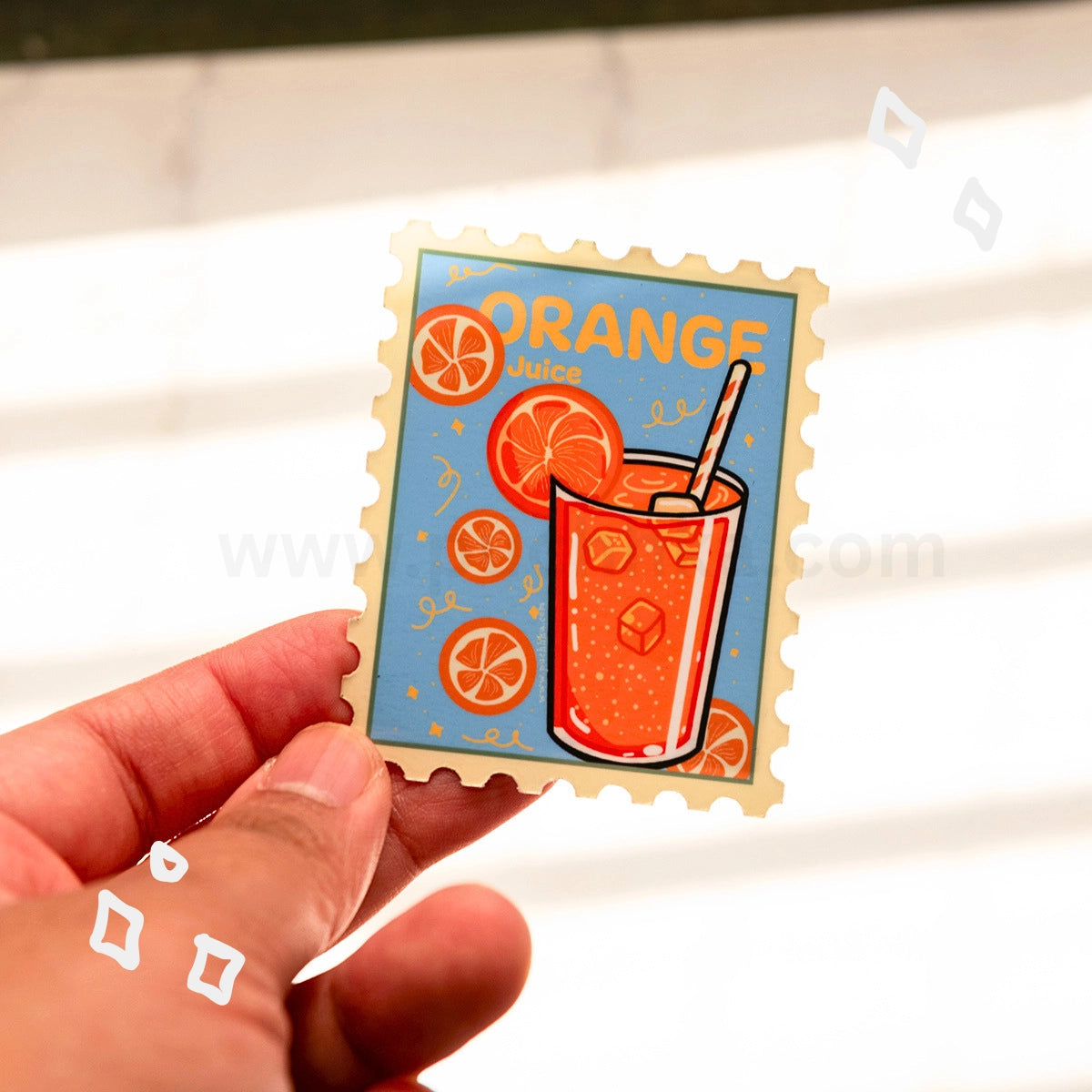 A hand holds a die-cut magnet featuring an orange-themed illustration with the word 'ORANGE' and a glass of orange juice, shown against a softly blurred light background.