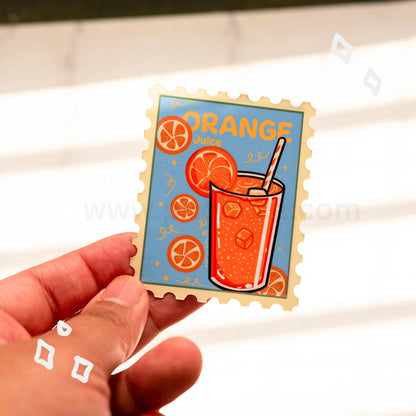A hand holds a die-cut magnet featuring an orange-themed illustration with the word 'ORANGE' and a glass of orange juice, shown against a softly blurred light background.