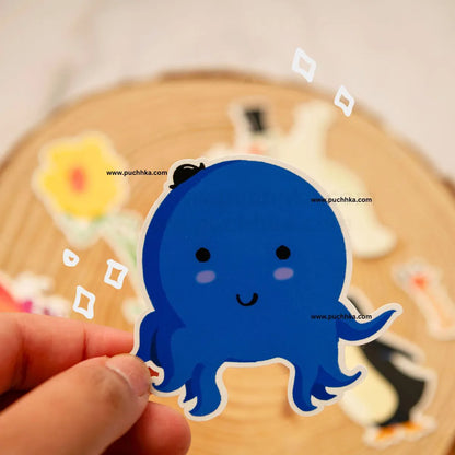 A hand holds a die-cut sticker of a cute blue octopus with a small black hat, set against a blurred background of other stickers on a wooden surface.