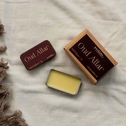 Solid perfume balm in a small metal tin shown open, alongside its kraft paper box and closed tin, labeled 'Oud Attar' by Puchhka, arranged on a light beige fabric background with a brown fringed textile at the side.