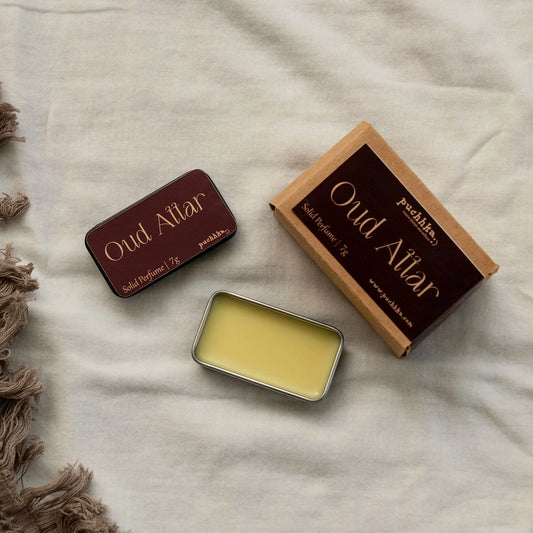 Solid perfume balm in a small metal tin shown open, alongside its kraft paper box and closed tin, labeled 'Oud Attar' by Puchhka, arranged on a light beige fabric background with a brown fringed textile at the side.