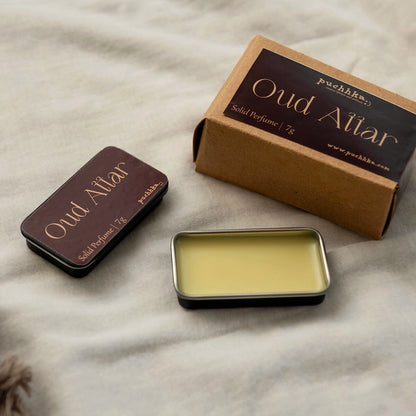 A product shot of Puchhka's Oud Attar solid perfume, showing the dark brown metal tin, its kraft paper packaging box, and the pale yellow wax inside, all arranged on a textured beige fabric background.