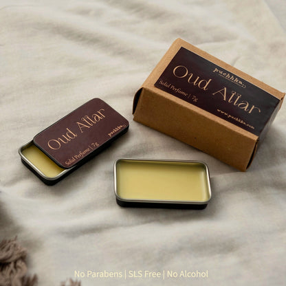 A small rectangular solid perfume tin shown open with pale yellow wax inside, alongside its kraft paper box labeled 'Oud Attar' and a second closed tin. The items are arranged on a light beige fabric background.