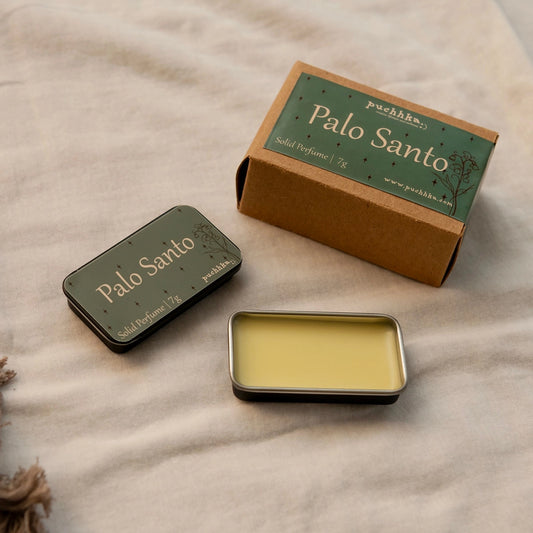 A small rectangular metal tin of Palo Santo solid perfume with pale yellow scented wax inside, shown beside its brown kraft box and closed tin on a light beige fabric surface.
