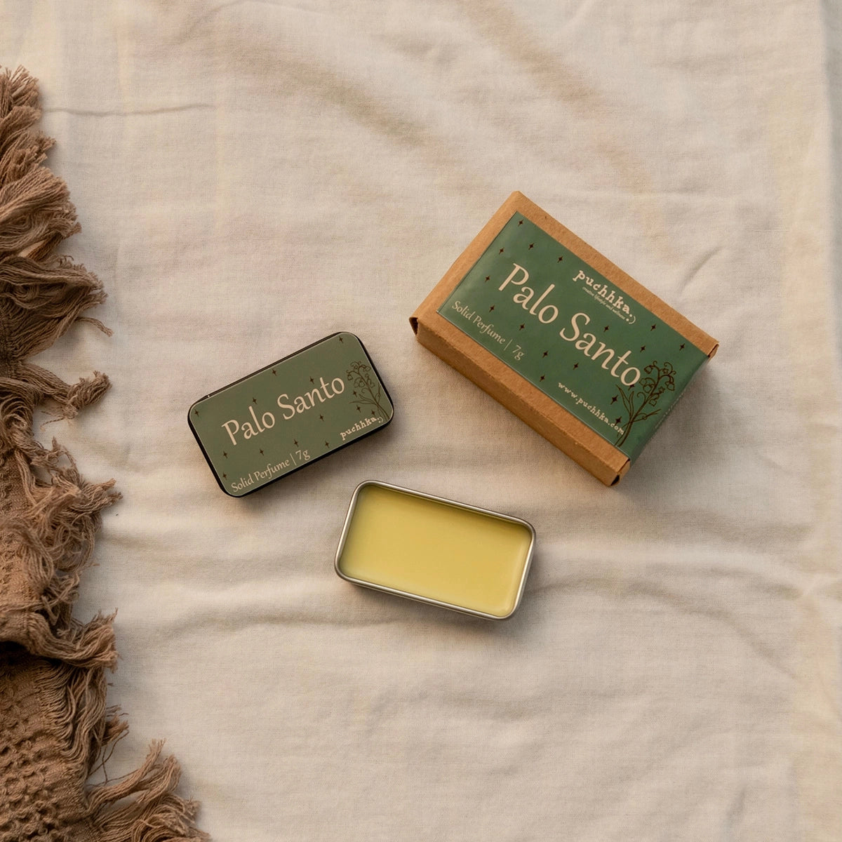 A styled flat-lay of Palo Santo solid fragrance wax products on a beige linen fabric background. The set includes a green metal tin lid, an open metal tin showing the pale yellow wax, and a brown kraft paper box with a green label.