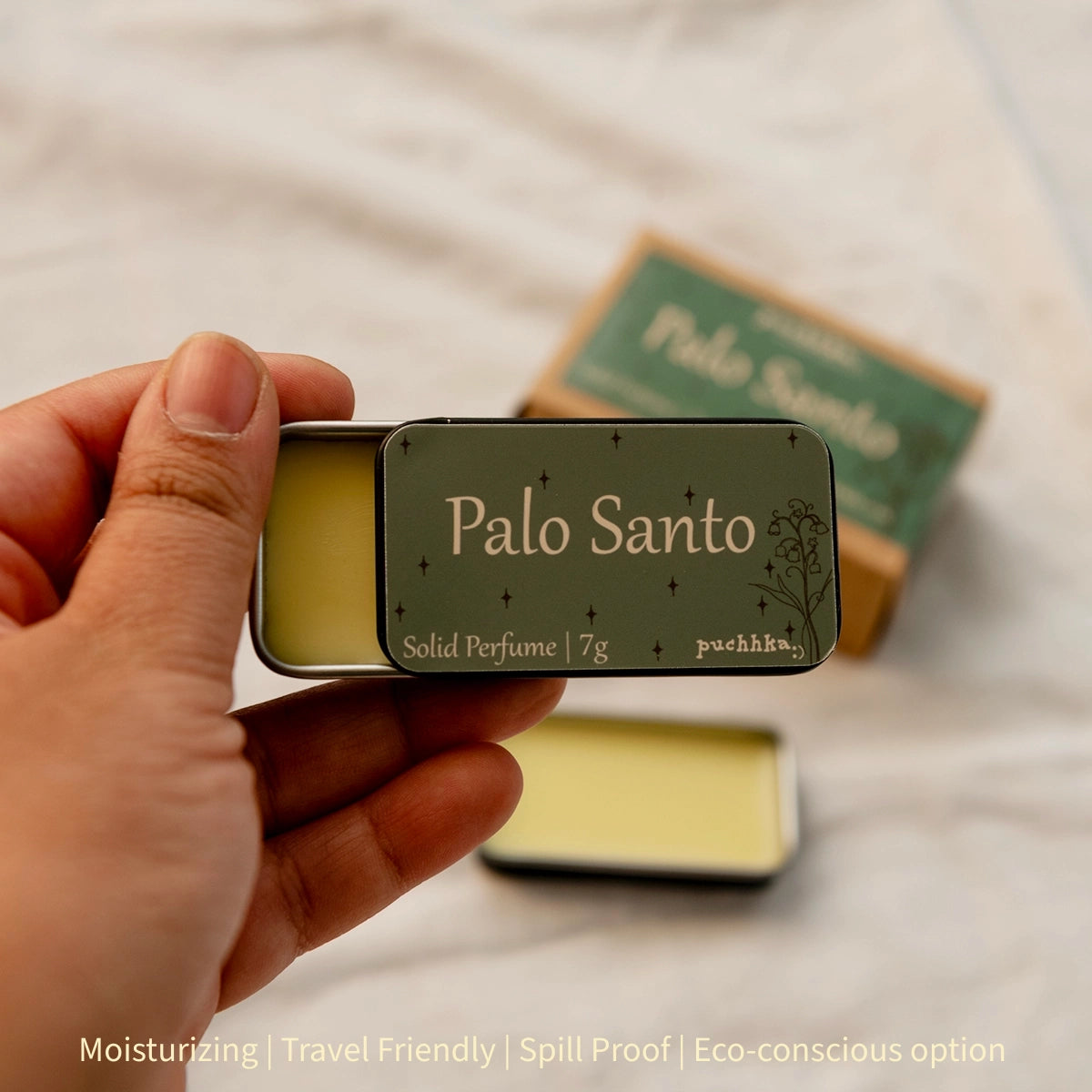 A hand holds an open metal tin of Palo Santo solid perfume, showing the pale yellow wax inside and the dark olive-green label. Another closed Palo Santo box sits blurred in the background on a light fabric surface.