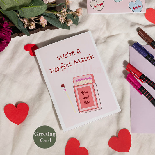 A square greeting card with the phrase 'We're a Perfect Match' and a pink matchbox illustration, styled on a cream fabric surface with red heart cutouts, flowers, markers, and a small 'Greeting Card' label.