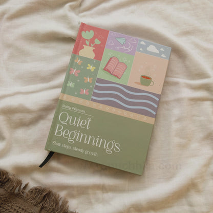 A Daily planner  'Quiet Beginnings' planner by Puchhka, featuring a pastel multicolor cover illustration, resting on a beige fabric background.